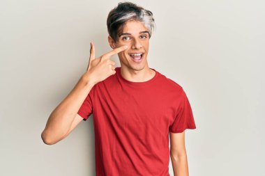 Latin asıllı genç bir adam, gündelik giysiler giyip el ve burnu işaret ediyor, neşeli bir şekilde gülümsüyor. güzellik konsepti 