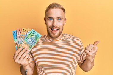 Young caucasian man holding australian dollars pointing thumb up to the side smiling happy with open mouth 