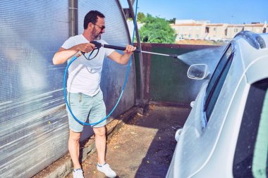 Orta yaşlı yakışıklı, günlük kıyafetler giyen ve mutlu gülümseyen güneş gözlüklü bir adam. Yüzünde gülümsemeyle ayakta durup su tabancası kullanarak araba yıkamak..