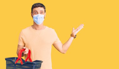 Alış veriş sepeti ve tıbbi maske takan yakışıklı genç adam zaferi mutlu bir gülümsemeyle ve kazanan yüz ifadesiyle kutluyor. 