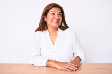Middle age brunette hispanic business woman wearing casual white shirt sitting on the table looking away to side with smile on face, natural expression. laughing confident. 