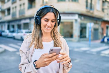 Beyaz kadın şehirde akıllı telefon ve kulaklık kullanarak mutlu bir şekilde gülümsüyor..