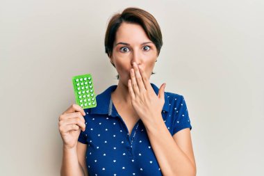 Kısa saçlı esmer bir kadın elinde doğum kontrol hapları tutarak ağzını eliyle kapatıyor, şok geçiriyor ve hata yapmaktan korkuyor. şaşırmış ifade 