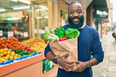 Sakallı Afrikalı Amerikalı bir adam elinde süpermarketten alınmış kağıt poşetler tutuyor.