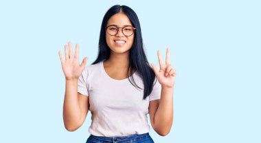 Genç, güzel Asyalı bir kız, günlük kıyafetler ve gözlük takıyor. Sekiz numaralı parmaklarıyla işaret ediyor ve gülümsüyor. Kendinden emin ve mutlu.. 