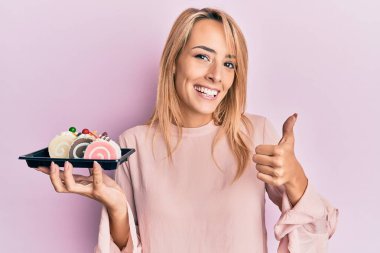 Güzel sarışın kadın elinde pasta şekerleri, gülümsüyor mutlu ve pozitif, başparmak yukarı, mükemmel ve onay işareti yapıyor. 