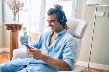 Genç İspanyol adam evde akıllı telefon ve kulaklık kullanarak mutlu bir şekilde gülüyor.