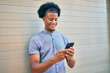 Genç Afrikalı Amerikalı adam şehirde akıllı telefon kullanarak mutlu bir şekilde gülümsüyor..