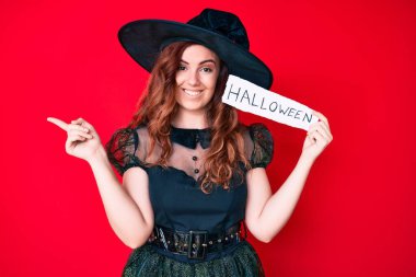 Cadı kostümü giymiş genç bir kadın Cadılar Bayramı mesajı içeren bir kağıt tutuyor. Gülümsüyor, mutlu bir şekilde işaret ediyor. 