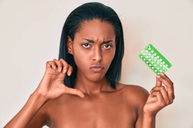 Genç Afro-Amerikalı kadın elinde kızgın yüzlü doğum kontrol hapları tutuyor, negatif işaret baş parmakları aşağıda, reddedilme konsepti 