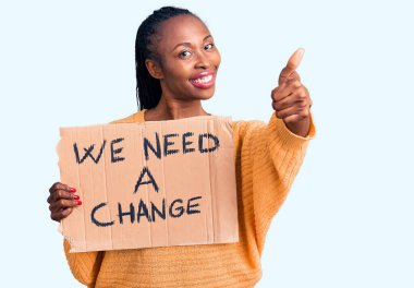 Genç Afro-Amerikan kadını elinde bir değişim pankartı tutarken mutlu ve olumlu bir şekilde gülümsemeli, başparmağını kaldırmalı ve mükemmel bir onay işareti koymalı. 