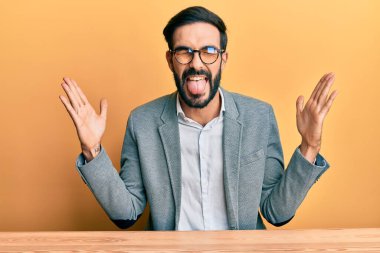 İş kıyafetleri giymiş, sakallı genç İspanyol bir adam masaya oturmuş deli gibi bağırıyor, bağırıyor, agresif bir ifade ve kollar havada. hüsran kavramı. 