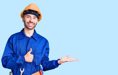 İspanyol asıllı genç adam işçi üniforması giyiyor, avuç içini gösteriyor ve baş parmaklarıyla iyi bir hareket yapıyor, mutlu ve neşeli gülümsüyor. 