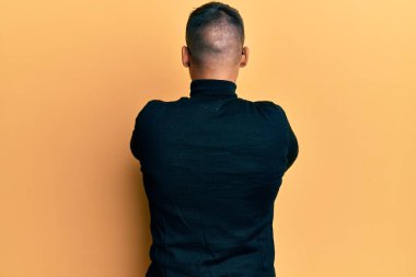 Balıkçı yaka kazak ve gözlük takan yakışıklı adam ters dönüp kollarını kavuşturarak başka tarafa bakıyor. 