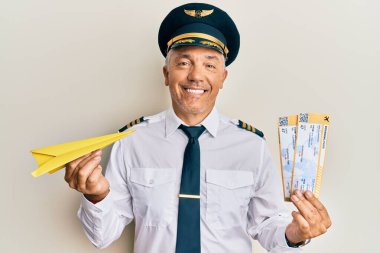 Elinde kağıt uçak ve biniş kartı olan yakışıklı, olgun bir pilot yüzünde mutlu ve havalı bir gülümsemeyle gülümsüyor. diş gösterme. 