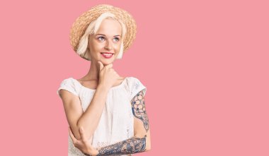 Yazlık şapkalı, elleri çenesinde, dövmeli soru soran genç sarışın kadın, düşünceli yüz ifadesi. Düşünceli bir yüzle gülümsüyor. şüphe kavramı. 