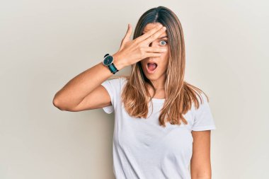 Esmer, beyaz tişörtlü genç bir kadın. Şoka girmiş, yüzünü ve gözlerini kapamış, parmaklarından korkuyla bakıyor. 