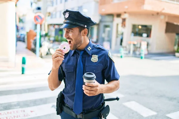 警察の制服を着た若いハンサムなパニック警官が笑顔で幸せ 町の通りでコーヒーを飲みながらドーナツを食べる — ストック写真