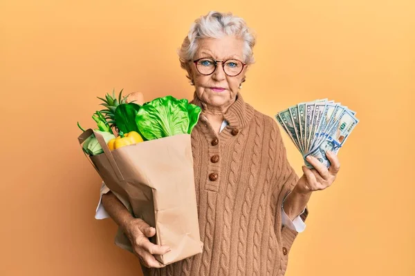 Senior grey-haired woman holding groceries and united states dollars banknotes relaxed with serious expression on face. simple and natural looking at the camera.