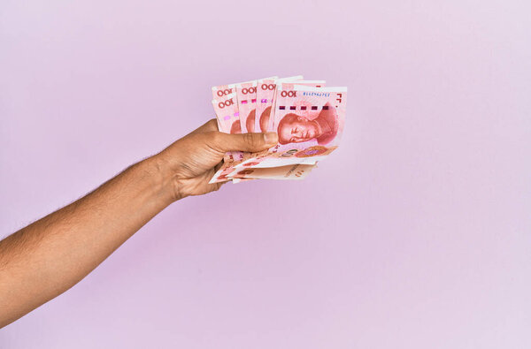 Hispanic hand holding 100 chinese yuan banknotes over isolated pink background.