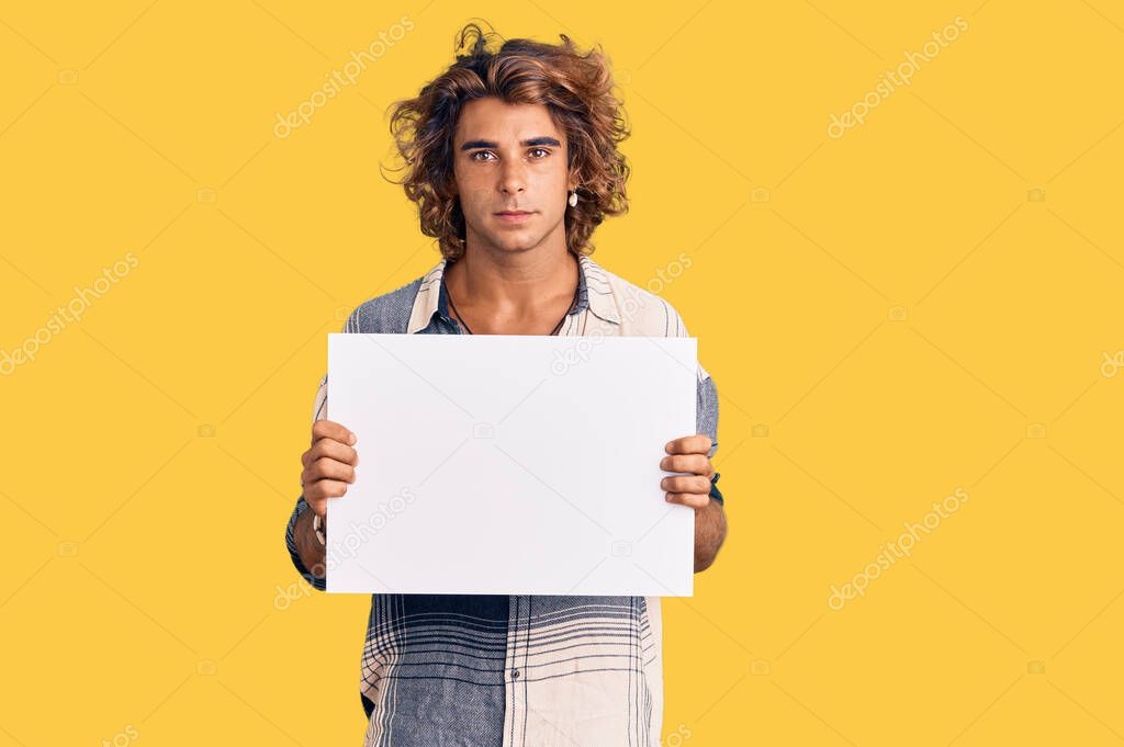Young hispanic man holding blank empty banner thinking attitude and sober expression looking self confident 