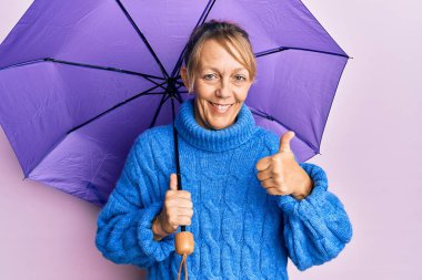 Orta yaşlı sarışın kadın elinde mor şemsiyeyle gülümsüyor mutlu ve pozitif, başparmağıyla mükemmel ve onay işareti yapıyor. 