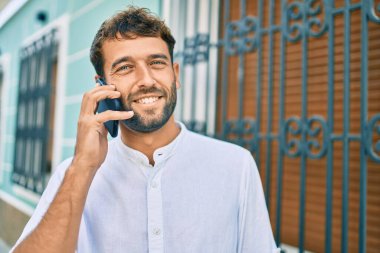 Güneşli bir günde sakallı, günlük beyaz gömlek giyen yakışıklı adam dışarıda mutlu bir şekilde gülümserken akıllı telefon kullanıyor.