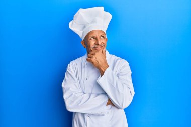 Orta yaşlı, gri saçlı, profesyonel aşçı üniforması giyen ve eli çenesinde şapkası olan soru soran, düşünceli bir adam. Düşünceli bir yüzle gülümsüyor. şüphe kavramı. 