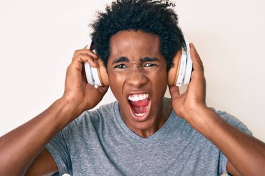 Handsome african american man with afro hair listening to music using headphones angry and mad screaming frustrated and furious, shouting with anger. rage and aggressive concept. 