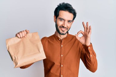 Elinde yiyecek tutan genç İspanyol adam parmaklarıyla imza atıyor, gülümseyerek mükemmel bir sembol oluşturuyor. 