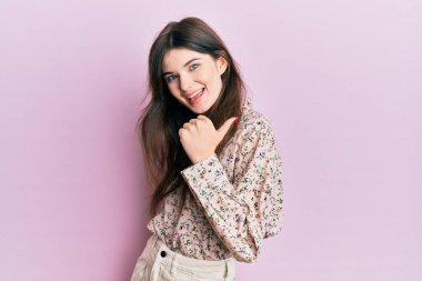 Zarif kıyafetler giyen, mutlu yüzüyle gülümseyen ve baş parmağıyla yüzünü gösteren güzel beyaz bir kız.. 