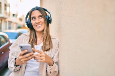 Beyaz kadın şehirde akıllı telefon ve kulaklık kullanarak mutlu bir şekilde gülümsüyor..