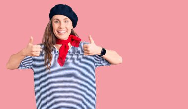 Genç, sarışın, Fransız beresi takan ve çizgili tişört giyen güzel bir kadın. Başparmağıyla gülümseyen ve mutlu bir şekilde olumlu jest yapıyor. Neşeli ifade ve kazanan jest. 