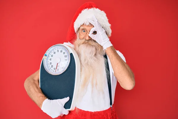 Old senior man on diet wearing traditional santa claus costume smiling happy doing ok sign with hand on eye looking through fingers