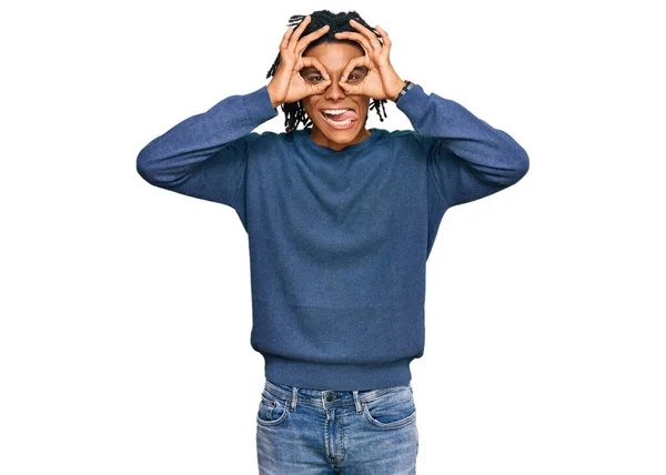 年轻的非洲裔美国人穿着宽松的冬季毛衣 做着像双筒望远镜一样的手势 伸出舌头 眼睛透过手指看过去 疯狂的表达 — 图库照片