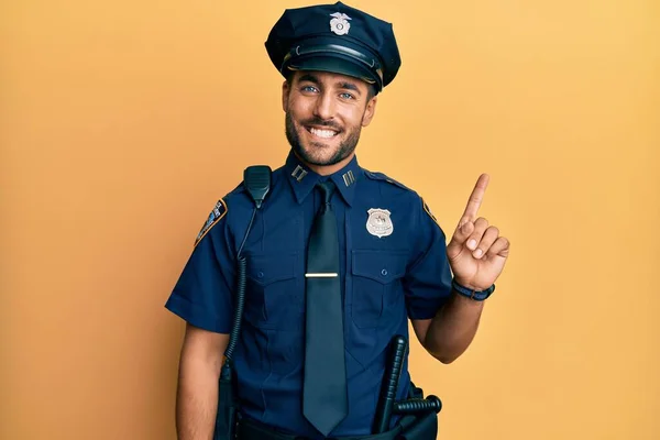 Handsome hispanic man wearing police uniform with a big smile on face, pointing with hand finger to the side looking at the camera.