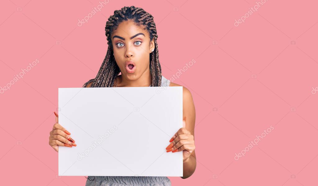Young african american woman with braids holding blank empty banner scared and amazed with open mouth for surprise, disbelief face 