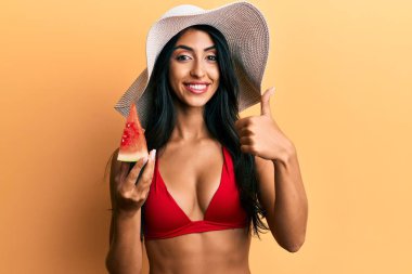 Güzel İspanyol kadın bikini giyip karpuz yiyor mutlu ve pozitif gülümsüyor, başparmağıyla mükemmel ve onay işareti yapıyor. 