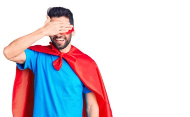 Sakallı, süper kahraman kostümü giyen, eli yüzü koyun gülen ve şaşırmak için gözlerini kapatan genç yakışıklı bir adam. kör kavram. 