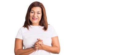 Middle age latin woman wearing casual white tshirt with hands together and crossed fingers smiling relaxed and cheerful. success and optimistic 