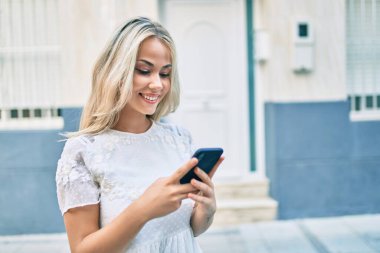 Beyaz kız şehrin caddesinde akıllı telefon kullanarak mutlu bir şekilde gülümsüyor..