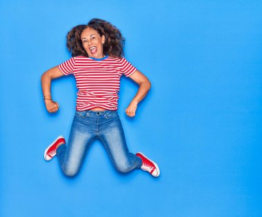 Orta yaşlı, güzel, İspanyol bir kadın. Günlük kıyafetler giyip mutlu gülümsüyor. İzole edilmiş mavi arka planda yüzünde gülümsemeyle zıplıyor