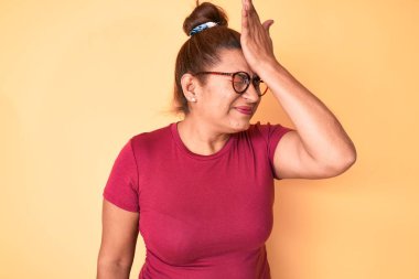Middle age brunette hispanic woman wearing casual clothes and glasses surprised with hand on head for mistake, remember error. forgot, bad memory concept. 