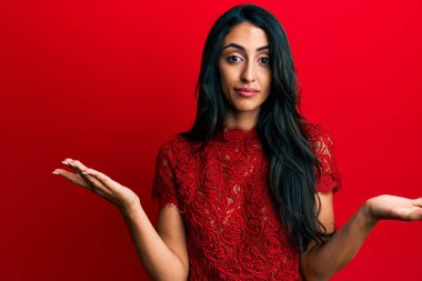 Güzel İspanyol kadın kırmızı arka planda zarif kıyafetler giyiyor. Hiçbir şeyden habersiz ve elleri ve kolları havada kafası karışmış bir ifade. şüphe kavramı. 