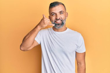 Orta yaşlı, beyaz tişört giyen, gülümseyen, el ve parmaklarıyla telefon hareketi yapan, telefonda konuşan yakışıklı bir adam. iletişim kavramları. 