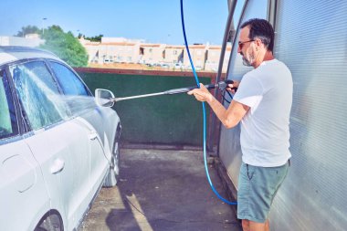 Orta yaşlı yakışıklı, günlük kıyafetler giyen ve mutlu gülümseyen güneş gözlüklü bir adam. Yüzünde gülümsemeyle ayakta durup su tabancası kullanarak araba yıkamak..