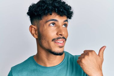 Gündelik giysiler giymiş genç Arap yakışıklı adam baş parmağını yana kaldırıyor ve ağzı açık gülümsüyor. 