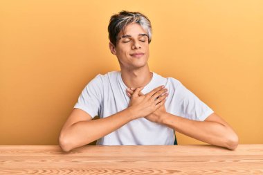 Gündelik giysiler giymiş genç İspanyol bir adam masanın üzerinde oturuyor elleri göğsünde gülümsüyor, gözleri kapalı ve yüzünde minnet dolu bir jest var. sağlık kavramı. 