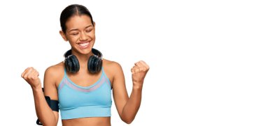 Spor kıyafetleri ve kulaklıklar takan güzel İspanyol bir kadın çok mutlu ve heyecanlı bir şekilde kollarını kaldırıp, gülümseyerek ve başarı için çığlık atarak kazanan bir jest yapıyor. kutlama konsepti. 