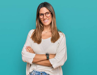 Sıradan kıyafetler ve gözlük takan, kollarını kavuşturup kameraya gülümseyen genç bir kadın. pozitif kişi. 